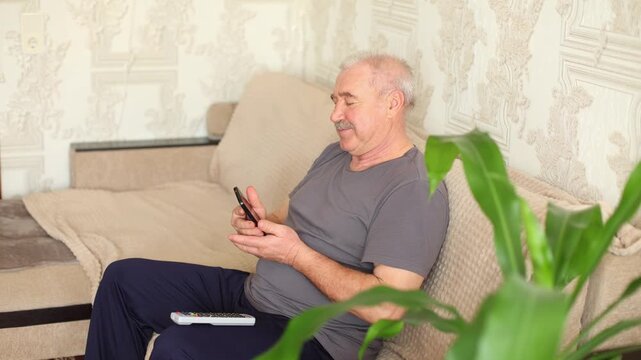 An older man with a mustache sits on a beige sofa, talking on a mobile phone, calm expression, patterned wallpaper, plant nearby.
