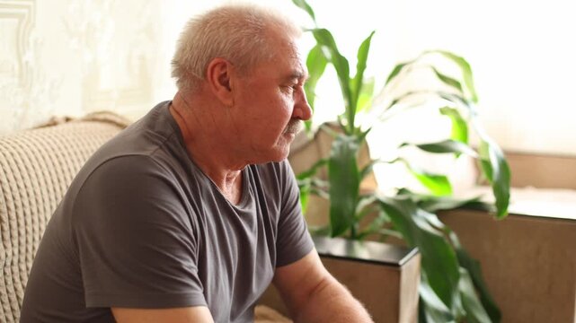 An older man with gray hair and a mustache sits on a sofa, holding a remote, gazing off screen, soft light, patterned wall, green plant in foreground.