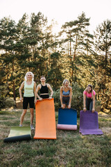 Smiling women standing with yoga mats outdoors in a park before starting group yoga. Fitness, wellness, friendship and healthy lifestyle concept.