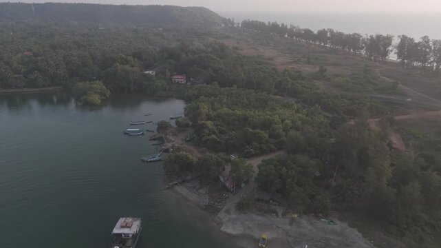Aerial drone view of Querim Beach at sunset from the Tiracol River, showing a silhouetted coastline with a row of trees, gentle waves, and a ferry crossing the calm river