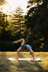 Fit woman performing one-legged wheel yoga pose outdoors, demonstrating advanced flexibility, core strength, and balance during sunset yoga practice in nature.