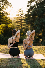 Two women practicing yoga shoulder stretch outdoors at sunset, kneeling on mats in a peaceful park. Wellness, flexibility and mindful movement concept.