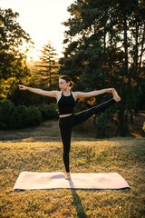 Woman practicing standing balance yoga pose while holding raised leg outdoors at sunset, focusing on flexibility, strength and concentration.
