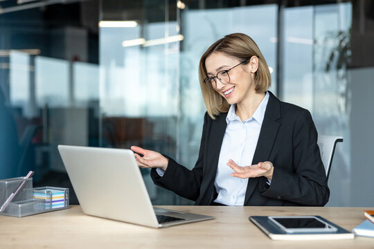 Young businesswoman with eyeglasses actively participating in an online video call or webinar using her laptop, presenting and smiling from a corporate office setting