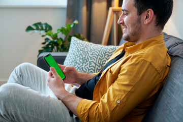 Young man sitting on sofa holding smartphone with green screen smiling and looking at device, relaxing at home with decorative plants in background