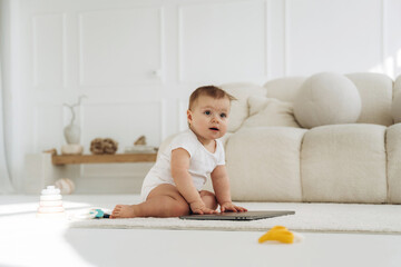 Fototapeta premium Baby in white onesie sits on soft carpet beside laptop and colorful toys in bright, modern living room with minimalist decor and natural light