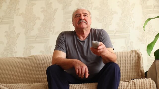 An older man with a mustache sits on a beige sofa, holding a remote, gazing thoughtfully, soft light, patterned wallpaper, and a houseplant frame the scene.