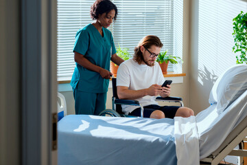 Young man with disability sitting in wheelchair using smartphone while Black woman nurse standing behind assisting in hospital room with sunlight streaming through blinds