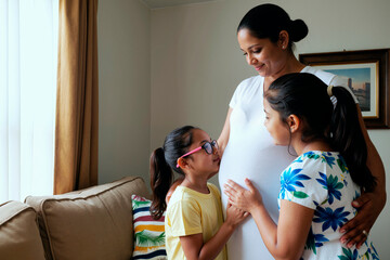 Pregnant woman standing in living room smiling while two girls, one wearing glasses, touching her belly and looking up at her with affection