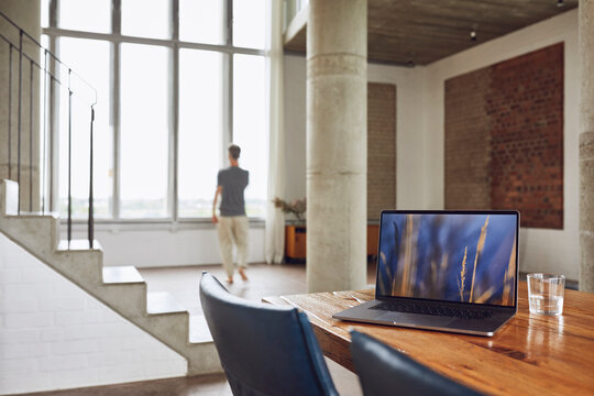 Laptop on wooden table in a loft flat with man at the window in background