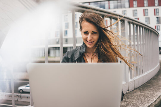 Portrait of smiling young woman with windswept hair using laptop on motorway bridge