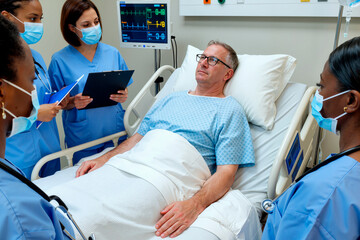 Adult man lying in hospital bed surrounded by multiethnic group of female nurses wearing medical masks discussing patient care and monitoring vital signs on screen