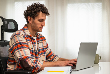 Confident and concentrated businessman sitting at the table while using laptop and working remotely at his cozy home.	