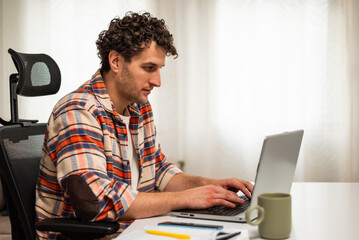 Focused businessman sitting at the table while using laptop and working remotely at his cozy home.	