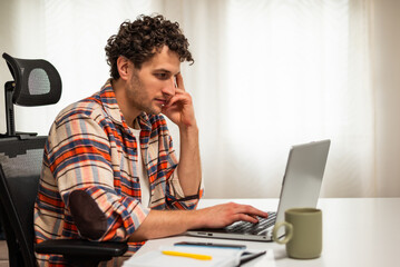 Successful young businessman thinking while using laptop and working remotely at his cozy home.