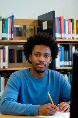 Portrait of young Black man sitting at desk in library writing with pencil, looking into camera, surrounded by bookshelves filled with academic books and study materials