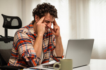 Despaired and exhausted young businessman having a headache while working on laptop at his home.	
