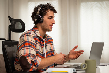 Young man with wireless headphones talking and gesturing during a video call on laptop while sitting in his cozy home.	