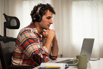 Modern young man wearing wireless headphones and using laptop while attending video conference. He is enjoying working remotely at his home office.	