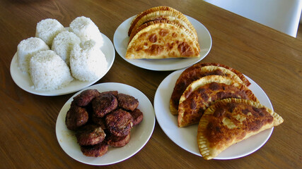 Traditional meal with rice balls fried pastries and meat patties. Home style food served on plates with rice balls empanadas and grilled meat on wooden table.