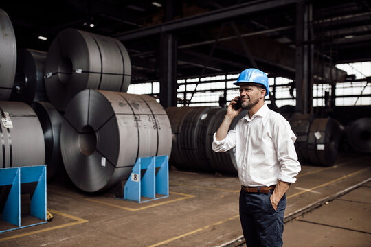 Smiling businessman with hand in pocket talking on smart phone while standing in industry