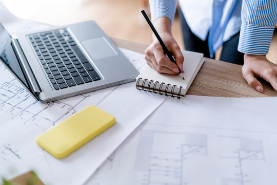 Close-up of woman in office taking notes with plan and laptop on table