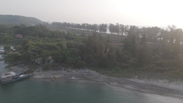Aerial drone view of Querim Beach at sunset from the Tiracol River, showing a silhouetted coastline with a row of trees, calm water, and a stationary ferry standing in the river