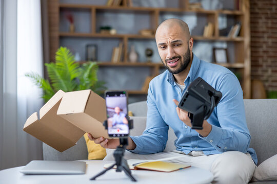 Man presenting a virtual reality headset to cellphone camera while live streaming video, expressing disappointment with the product during an unboxing and tech review for his online audience