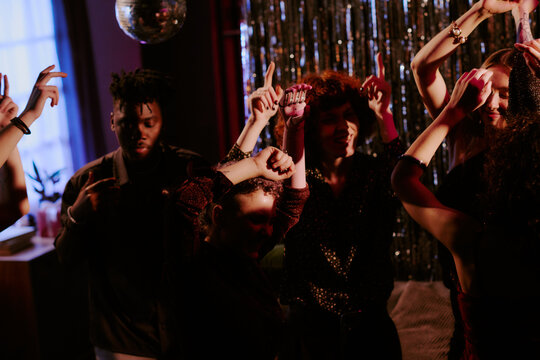 Group of young adult multiethnic men and women dancing together at party, raising arms and moving to music in dimly lit room with shimmering backdrop and disco ball overhead