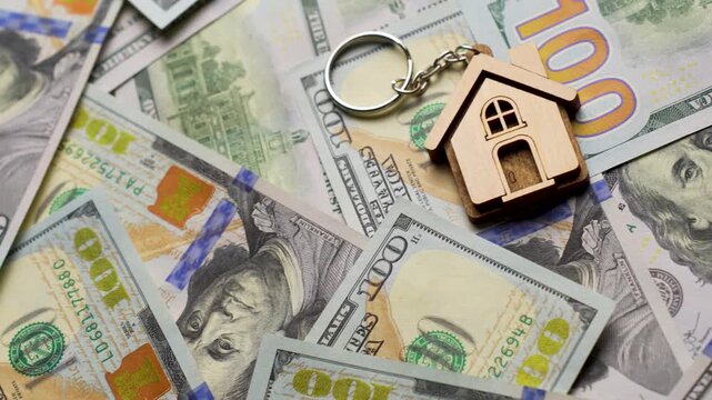 Property Investment and Home Ownership Concept. A wooden house shaped keychain with silver keys lying on a background of scattered dollar bills