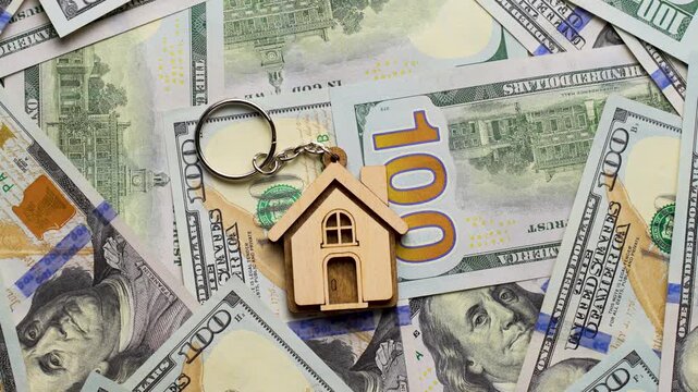 Property Investment and Home Ownership Concept. A wooden house shaped keychain with silver keys lying on a background of scattered dollar bills