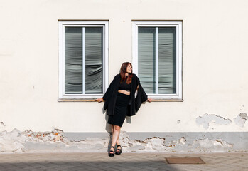 Stylish woman in black outfit poses confidently against a textured wall with windows, showcasing modern fashion and urban lifestyle in a bright, sunlit environment