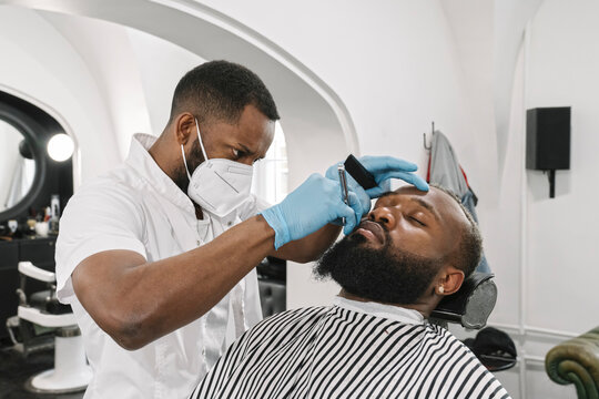 Man with full beard getting a shave, barber wearing surgical mask and gloves