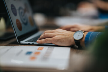A person types on a laptop at a desk in a professional office setting with a wristwatch and charts.