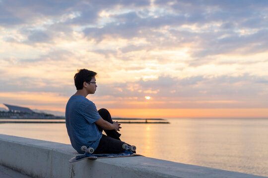 Young Chinese man with skateboard sitting on wall at the beach