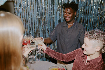 Naklejka premium Group of young adults including Black man and Caucasian women clinking glasses and smiling during celebration, standing together in front of metallic streamers at festive event