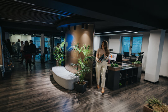 A professional woman holds a folder while walking in a contemporary office lobby. The scene shows coworkers gathered in the background near glass walls and modern workstations.