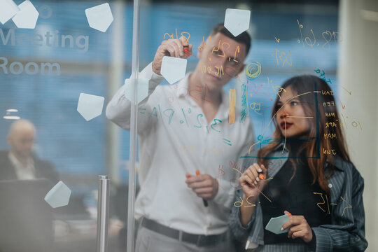 Two business colleagues brainstorm on a glass whiteboard in a modern office. The scene captures collaboration, ideas, and problem solving.