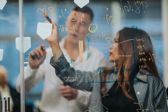 Two business colleagues collaborate on ideas at a glass board filled with colorful equations. A planning session unfolds as they write and discuss strategies.