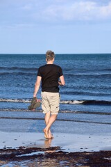 Mann geht mit Badelatschen in der Hand am Strand spazieren
