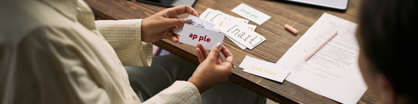 Caucasian woman tutor holding flashcard with word apple, teaching young student during lesson at desk with educational materials visible hands and learning cards on wooden table