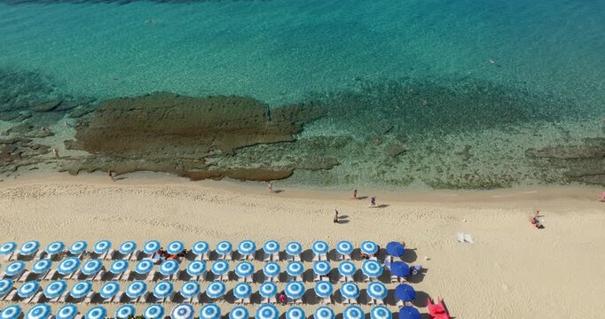 Aerial view of a sandy beach featuring neatly arranged rows of beach umbrellas. The shoreline reveals clear turquoise water with visible rocky formations under the water surface. Summer morning.