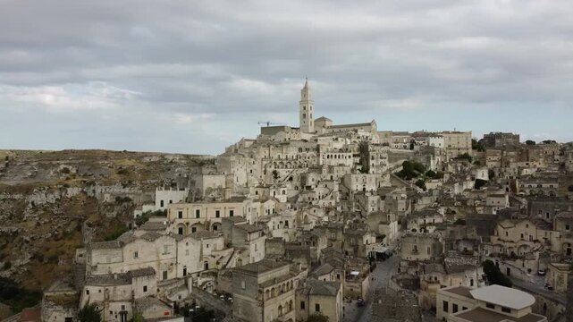 Establishing aerial drone shot of Matera, Italy, captured in summer under a cloudy sky. Panoramic footage revealing the iconic Sassi cave dwellings and historic urban landscape.