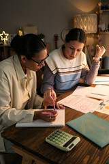 Obraz premium Vertical of young adult Caucasian woman tutoring young adult Black woman at desk, both writing in notebooks and reviewing documents together, calculator and stationery on table, focused on lesson