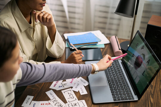 Young Caucasian girl sitting with young Black woman tutor using laptop for online lesson, both pointing at screen with phonetic cards and notebooks on desk, engaging in interactive learning