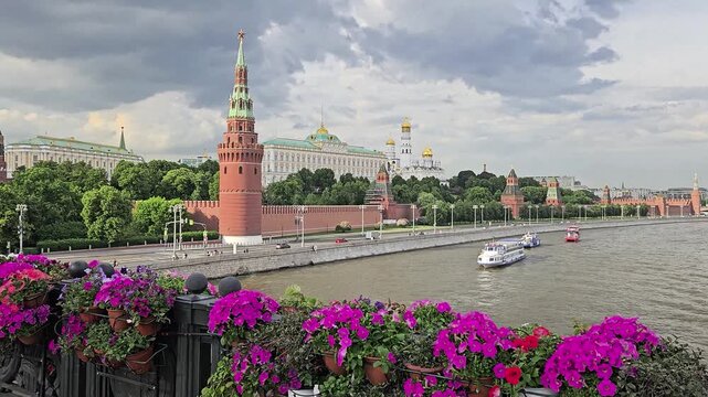 Boat is traveling down a river near a city Moscow. The river is lined with Kremlin wall, buildings and trees. The bridge has a row of potted plants with flowers