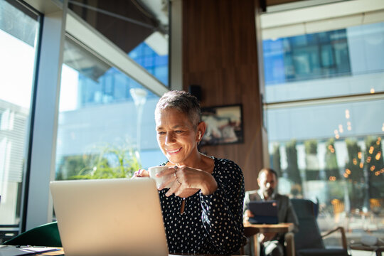 Smiling mature woman with earbuds working on laptop in modern cafe
