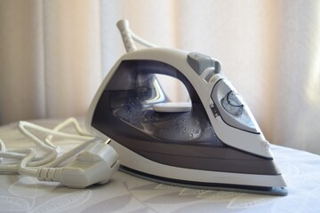 Close-up of an iron with a cord on an ironing board against the background of curtains in a room