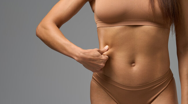 Close-up of woman in beige underwear pinching skin on side of stomach in studio light.