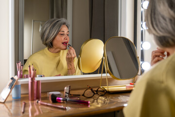 Elegant senior woman with short grey hair applying red lipstick, looking at her reflection in a lighted vanity mirror